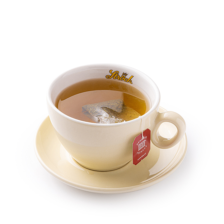 Eine beigefarbene Tasse mit einem Teebeutel und einem roten Anhänger, gefüllt mit Tee, steht auf einer passenden Untertasse vor einem weißen Hintergrund. Die Tasse ist mit einem goldenen Text beschriftet.