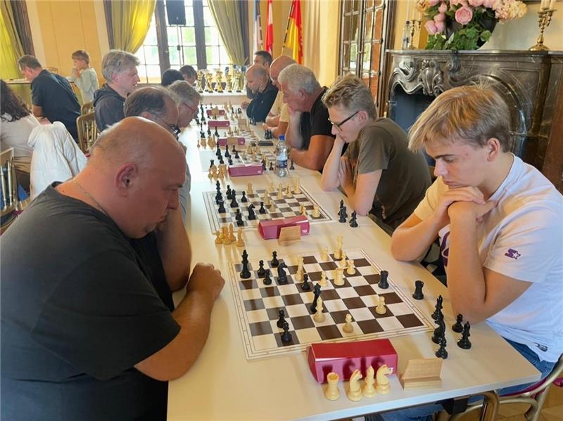 Mehrere Personen sitzen in zwei Reihen an einem langen Tisch und spielen konzentriert Schach in einem gut beleuchteten Raum mit großen Fenstern, dekorativen Blumen und einem verschnörkelten Kamin im Hintergrund.