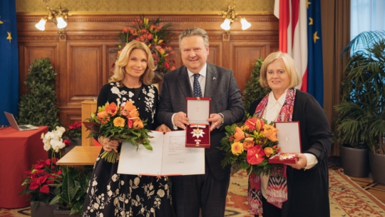 Drei Personen stehen lächelnd in einem Raum. Der Mann in der Mitte hält eine Medaille in einer Schachtel, flankiert von zwei Frauen mit Blumen und Urkunden. Eleganter holzgetäfelter Raum mit Fahnen und Blumenschmuck im Hintergrund.