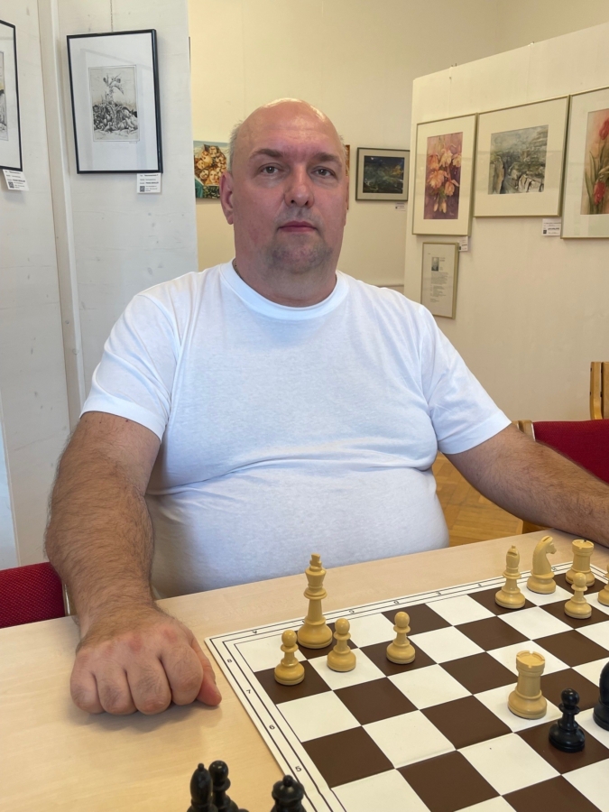 Ein Mann in einem weißen T-Shirt sitzt an einem Tisch vor einem Schachbrett und ist bereit, Schach zu spielen. Er befindet sich in einem Raum, in dem gerahmte Kunstwerke an den Wänden hinter ihm hängen.