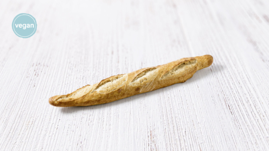 Ein einzelnes Baguette mit einer goldenen Kruste liegt auf einer hellen Holzoberfläche. Auf einem runden blauen Etikett in der linken oberen Ecke steht "vegan".