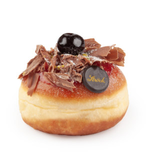 Ein runder, mit Gelee gefüllter Donut mit Schokoladenraspeln, einer glänzenden Kirsche und einer kleinen Schokoladenscheibe mit der Aufschrift Ströck auf weißem Hintergrund.