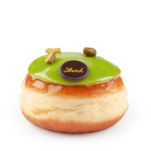 Ein runder, fluffiger Donut mit grüner, glänzender Glasur, ein paar Pistazienstücken und einer kleinen Schokoladenscheibe mit goldener Schrift. Der Donut befindet sich auf einem schlichten weißen Hintergrund.