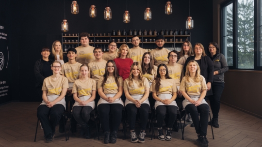 Eine Gruppe von sechzehn Personen, meist junge Erwachsene in passenden beigen Hemden, posiert in einem Innenraum auf Stühlen und im Stehen. Drei Erwachsene in dunklerer Kleidung stehen dahinter. Im Hintergrund befinden sich Regale mit Flaschen und Hängelampen.