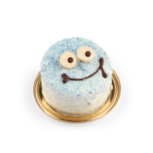 Ein kleiner, runder Kuchen, der wie ein lächelndes Gesicht mit blauem und weißem Zuckerguss, zwei großen Süßigkeitenaugen und einem Schokoladenlächeln dekoriert ist und auf einem goldenen Tablett steht.