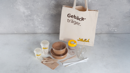 Hinter Kaffeebechern, Schüsseln, einem Glas mit gelbem Deckel, einem Glas mit Strohhalm, Servietten, Holzbesteck und Papierstrohhalmen steht auf einer hellen Fläche eine wiederverwendbare Tragetasche mit dem Schriftzug Gebäck-träger.