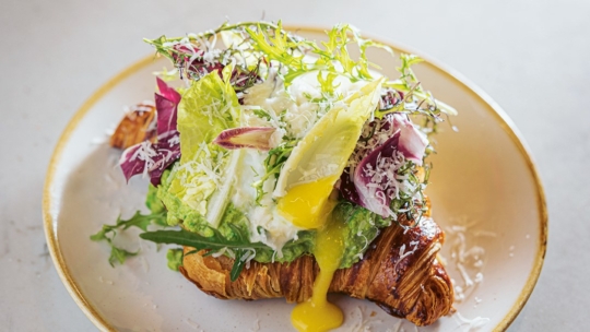 Ein Croissant, belegt mit Blattgemüse, geriebenem Käse, Avocado und einem pochierten Ei mit herausquellendem Eigelb, serviert auf einem beigen Teller.