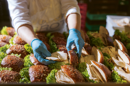 Eine Person mit blauen Handschuhen stellt Sandwiches mit Brotscheiben, Truthahn und Salat zusammen und legt sie auf einen Tisch, auf dem viele ähnliche Sandwiches liegen.