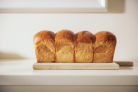 Ein frischer Laib goldbraunen Brotes liegt auf einem hölzernen Schneidebrett auf einer weißen Arbeitsplatte, die von sanftem Tageslicht erhellt wird.