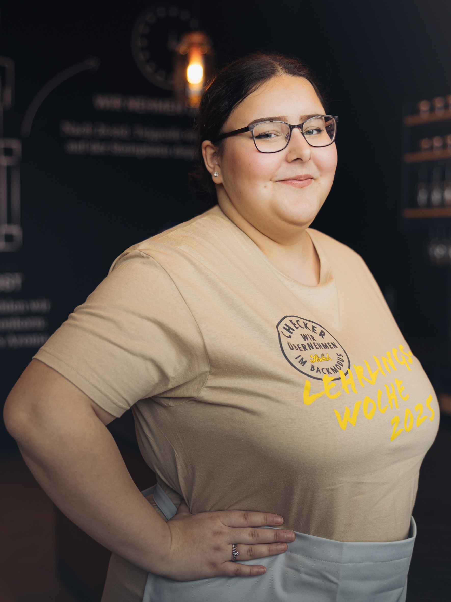 Eine Frau mit Brille lächelt selbstbewusst und stützt eine Hand in die Hüfte. Sie trägt ein beiges Hemd mit dem Aufdruck "Lehrlings Woche 2023" und eine helle Schürze. Im Hintergrund ist eine Schrift auf einer dunklen Wand leicht verschwommen.