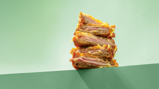 Ein Stapel von vier gegrillten Schinken-Käse-Sandwiches ist ordentlich auf einer grünen Fläche aufgestapelt, wobei geschmolzener Käse und Schichten von Schinken vor einem hellgrünen Hintergrund zu sehen sind.