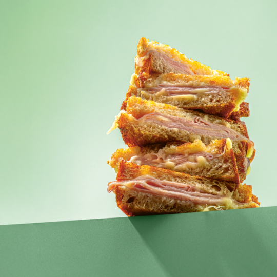 Ein Stapel von vier Schinken-Käse-Sandwiches mit getoastetem Brot wird auf einer grünen Fläche vor einem hellgrünen Hintergrund balanciert.