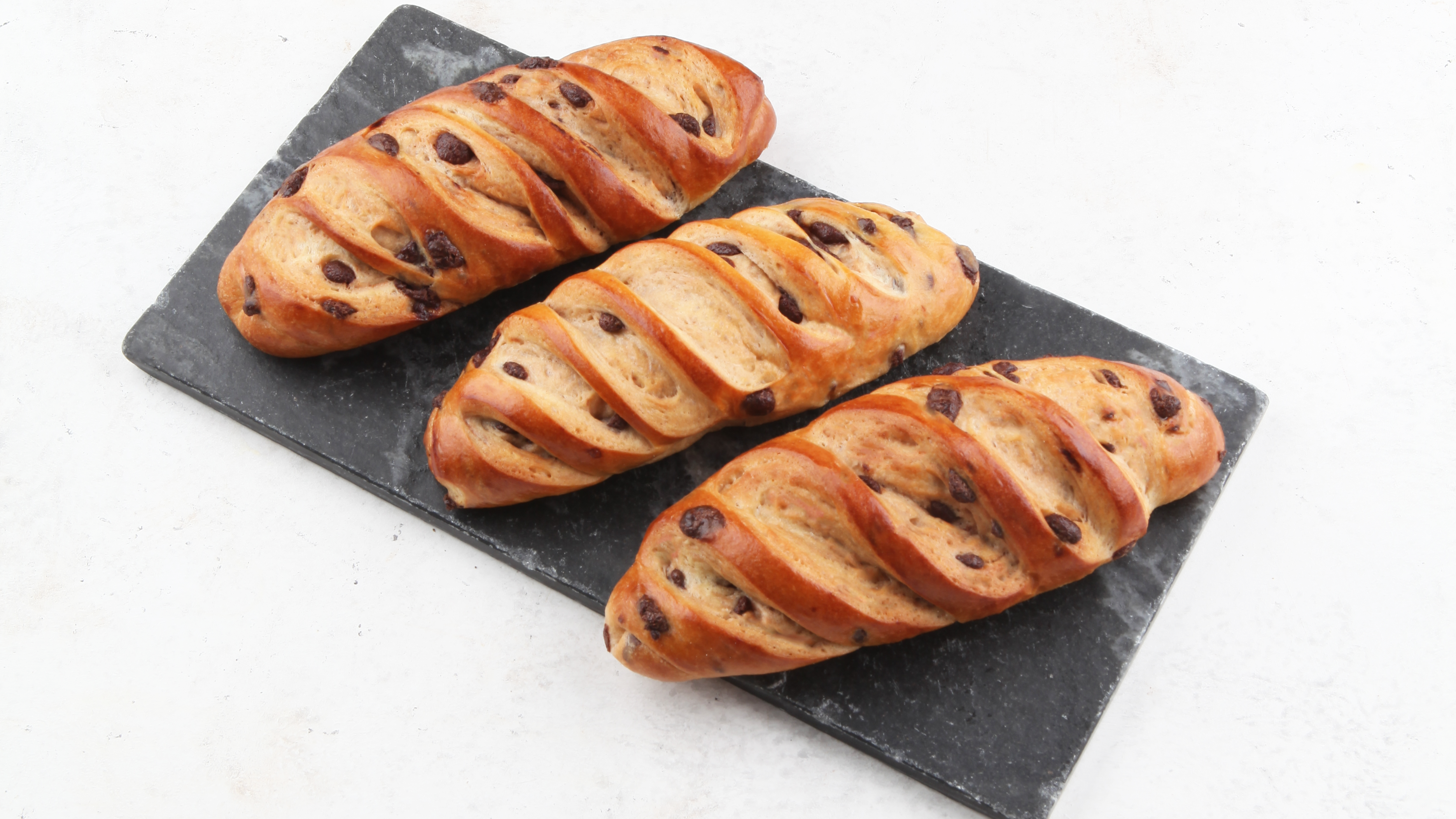 Drei goldbraune, geflochtene Brote mit Schokoladenstückchen sind auf einer rechteckigen schwarzen Schiefertafel vor einem weißen Hintergrund angeordnet.