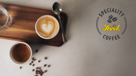Zwei Kaffeetassen auf einem Holztablett mit Löffel, eine davon mit herzförmiger Latte Art, daneben verstreute Kaffeebohnen. Rechts das Logo von SPECIALITY COFFEE Ströck mit einer Grafik von Kaffeebohnen.