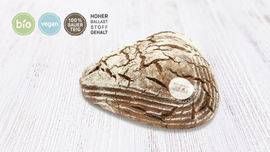 Ein rustikaler, dreieckiger Brotlaib mit einer knusprigen, rissigen Oberfläche liegt auf einer hellen Holzfläche. Auf den vier Etiketten darüber steht: bio, vegan, 100% Sauerteig und hoher Ballaststoffgehalt.