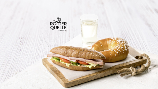 Ein Sandwich mit Schinken, Salat und Tomate, ein rundes Gebäck mit grobem Zucker und ein Glas Wasser sind auf einem Holzbrett angeordnet. Im Hintergrund ist das Logo der Römerquelle zu sehen.