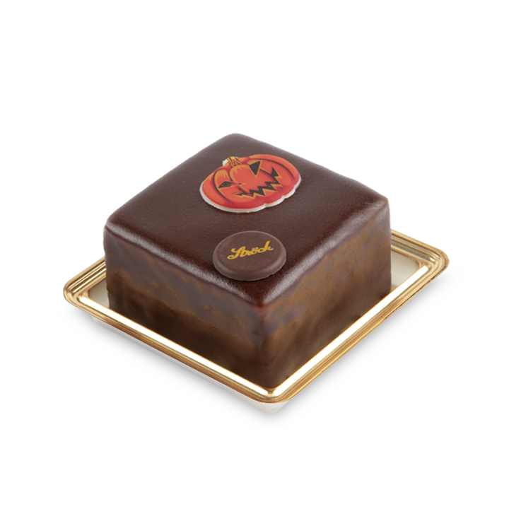 Ein quadratischer Schokoladenkuchen, verziert mit einer Kürbisdekoration und einer kleinen Scheibe mit dem Ströck-Logo, serviert auf einem goldgerahmten Tablett.