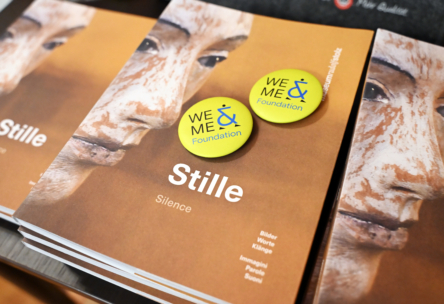 Several copies of a book titled “Stille (Silence)” with a close-up photo of a carved face on the cover. Two yellow “WE & ME Foundation” buttons rest on top of the books.