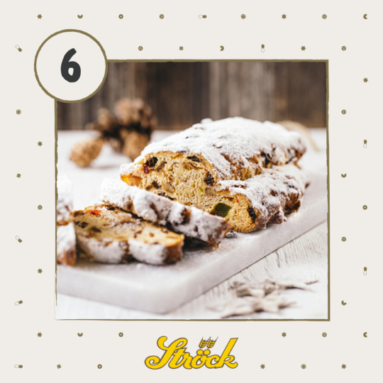 A loaf of stollen bread dusted with powdered sugar is sliced on a marble board. The background is soft-focused, with pine cones and a wooden surface, and the number 6 appears in the top left corner. The Ströck logo is at the bottom.