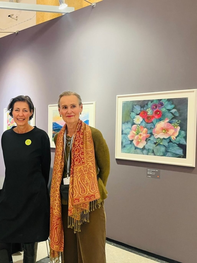 Two women stand smiling in front of a grey wall displaying colorful floral paintings in frames at an art exhibition. One woman wears a black dress, the other has a patterned scarf over a green outfit.