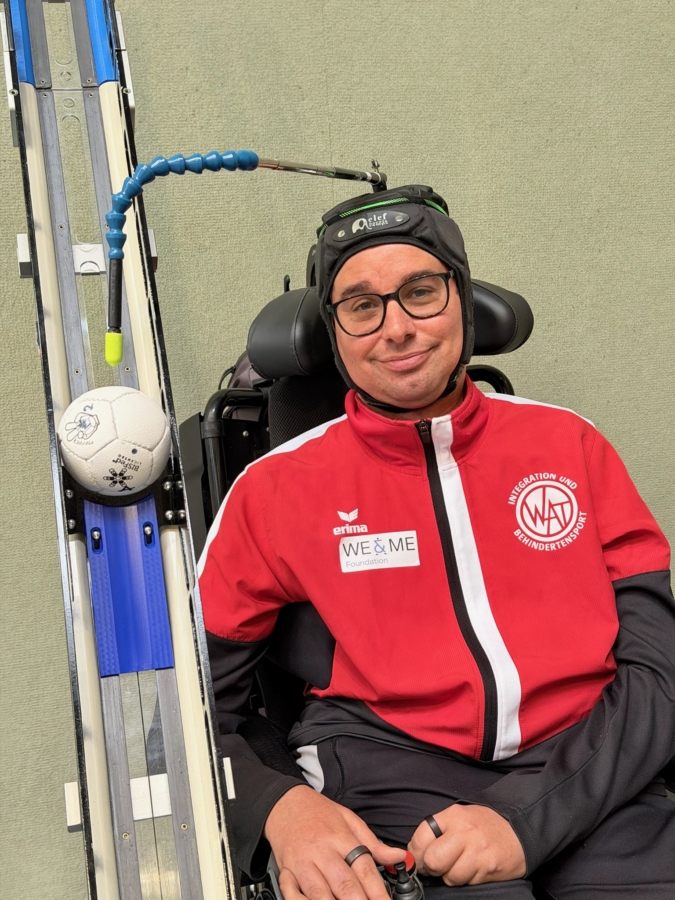A man wearing glasses and a red sports jacket sits in a wheelchair, using a head pointer to control a ramp holding a soccer ball, indicating participation in an adaptive sports activity.