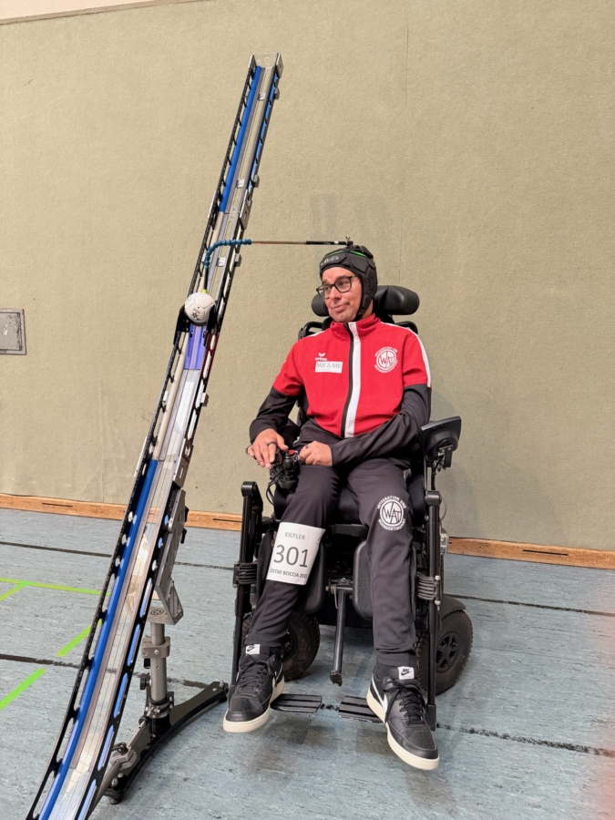 A man in a wheelchair wears sports gear and glasses. He is positioned next to a tall ramp holding a ball, likely for a boccia game. The scene takes place indoors on a gym floor.