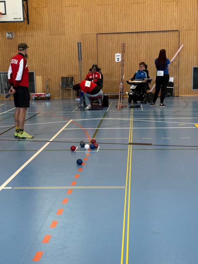 Eine Gruppe von Sportlern, darunter zwei Rollstuhlfahrer, nimmt an einem Boccia-Spiel in einer Halle teil. Die Bälle sind auf dem Spielfeld verstreut, und ein Schiedsrichter steht in der Nähe. Der Schauplatz ist eine Turnhalle mit Holzwänden und blauem Bodenbelag.