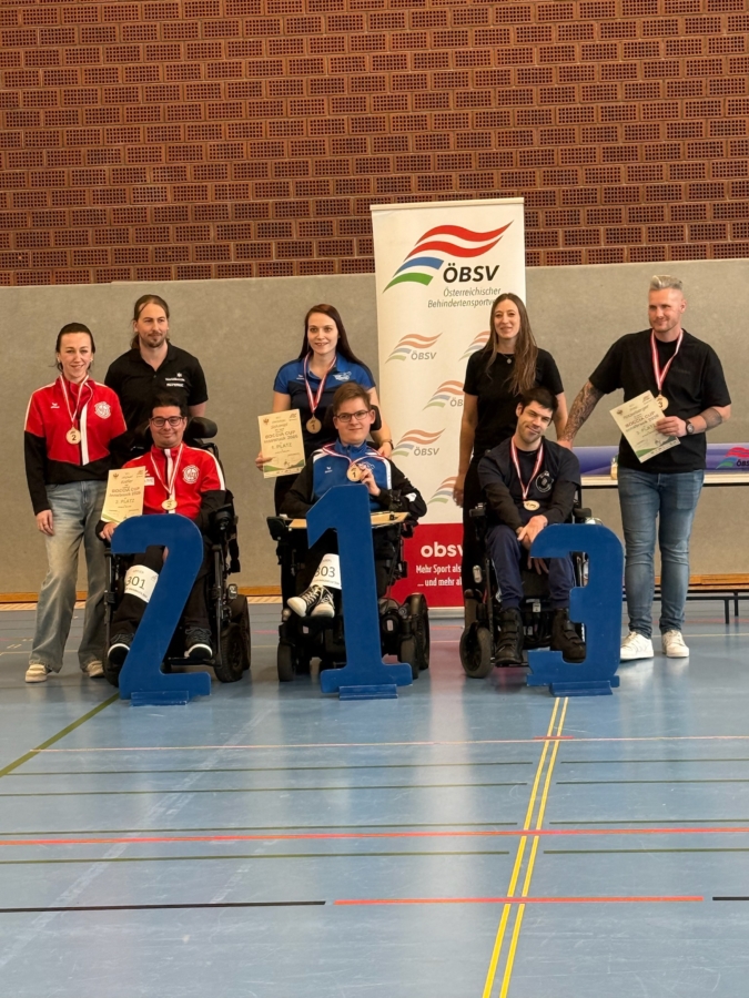 Acht Personen posieren in einer Sporthalle mit Medaillen und Urkunden, drei davon in Rollstühlen mit den großen blauen Zahlen 2, 1 und 3, vor einem ÖBSV-Banner auf dem Boden einer Sporthalle.