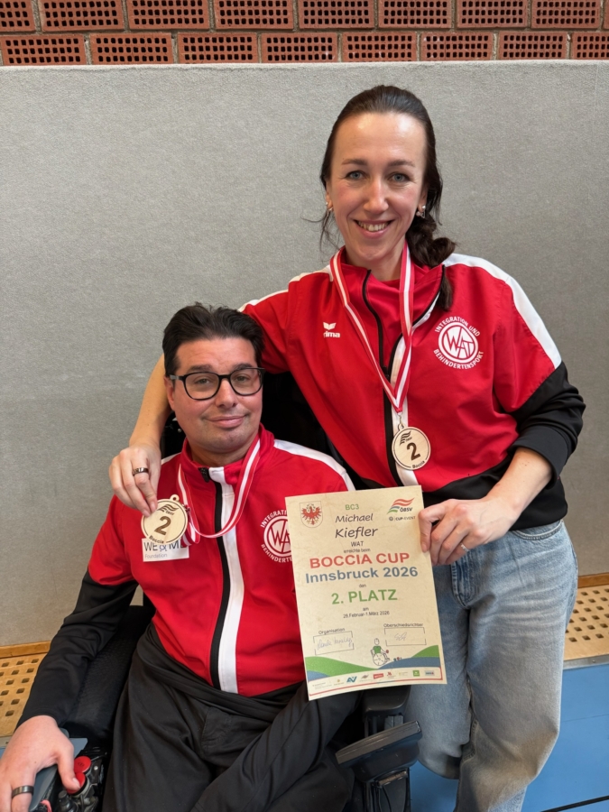 Zwei lächelnde Athleten in passenden rot-schwarzen Jacken posieren mit silbernen Medaillen um ihre Hälse. Einer sitzt in einem Rollstuhl. Sie halten eine Urkunde mit der Aufschrift "Boccia Cup Innsbruck 2026, 2. Platz".