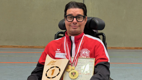 A man in a wheelchair, wearing glasses and a red sports jacket, smiles while holding a trophy, medals, and a certificate in a gymnasium.