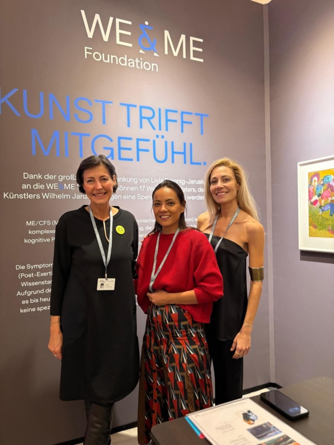 Three women stand smiling in front of a WE & ME Foundation display with German text on a wall. They wear conference badges and professional attire, and a table with documents and a binder is visible in the foreground.