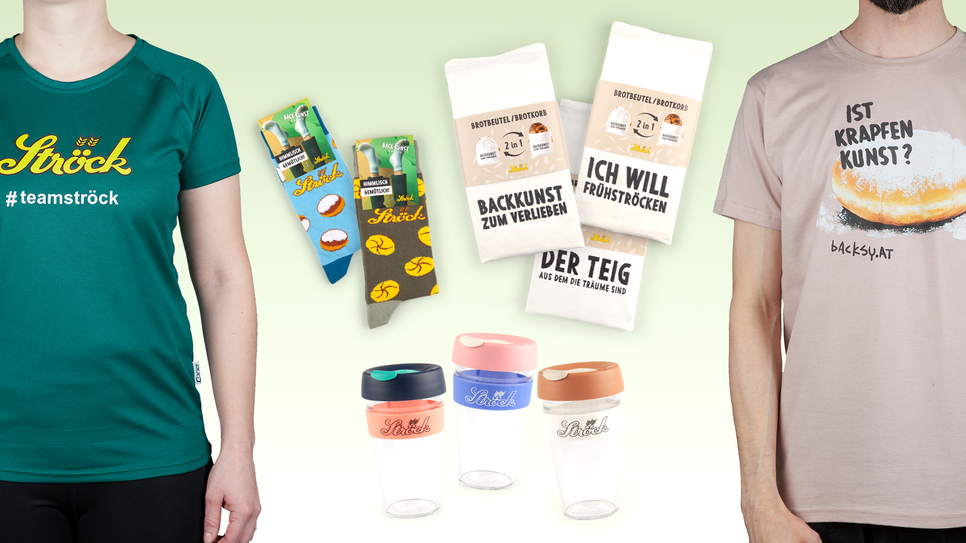 Eine Warenauslage mit zwei T-Shirts, bunten Socken, zwei weißen Küchentüchern mit deutschem Text und vier wiederverwendbaren Bechern zum Mitnehmen mit verschiedenfarbigen Deckeln, die alle ein Bäckereimotiv haben.