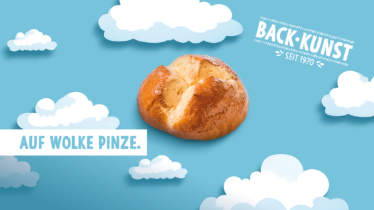Ein rundes, goldenes Brötchen schwebt zwischen illustrierten weißen Wolken auf blauem Hintergrund. Der Text lautet "AUF WOLKE PINZE." und "BACK-KUNST SEIT 1970." und zelebriert Bäckerei-Tradition mit einem Hauch von Laune.