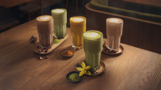Vier hohe Latte-Gläser in verschiedenen Geschmacksrichtungen - Schokolade, Kurkuma, Matcha und Chai - mit herzförmigen Latte-Kunstwerken stehen auf einem Holztisch mit den passenden Zutaten und Gewürzen daneben.