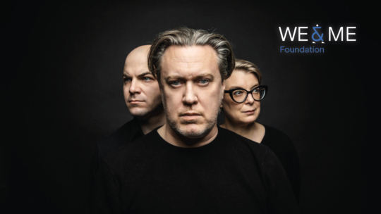 Drei ernste Erwachsene in schwarzer Kleidung stehen vor einem dunklen Hintergrund. Der Mann vorne hat graues Haar, links steht ein Mann mit rasiertem Kopf und rechts eine Frau mit Brille. Oben rechts steht WE & ME Foundation geschrieben.