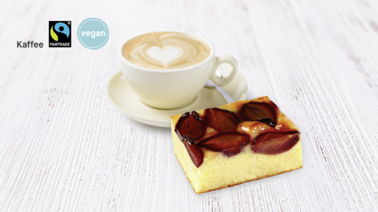 Eine Tasse Kaffee mit Latte Art auf einer Untertasse und ein Stück Pflaumenkuchen auf einer weißen Holzfläche. In der oberen linken Ecke sind Fairtrade- und Vegan-Labels sowie das Wort Kaffee zu sehen.