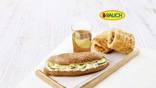 Ein Sandwich mit Salat und Gurke, ein Glas Apfelsaft und zwei Backwaren auf einem Holzbrett mit dem RAUCH-Logo im Hintergrund. Der Hintergrund ist hell und rustikal.
