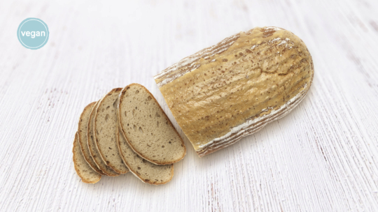 Ein in Scheiben geschnittenes Schwarzbrot auf einer weißen Holzfläche, mit drei aufgefächerten Scheiben davor. In der Ecke steht ein blaues, rundes Etikett mit der Aufschrift "vegan".