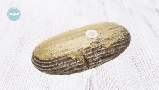 Ein runder, brauner Brotlaib mit einer leicht bestäubten Kruste und einer hellbraunen Glasur auf der Oberseite, der auf einer weißen Holzfläche liegt. In der oberen linken Ecke befindet sich ein blaues veganes Etikett.