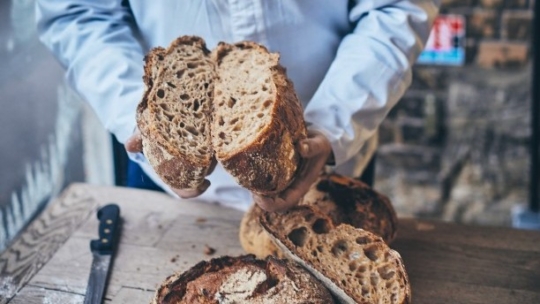 Eine Person in einem hellblauen Hemd hält einen rustikalen, runden Brotlaib in der Hand und spaltet ihn, so dass sein strukturiertes Inneres sichtbar wird. Ein Messer und weitere Brote liegen auf einem Holztisch.
