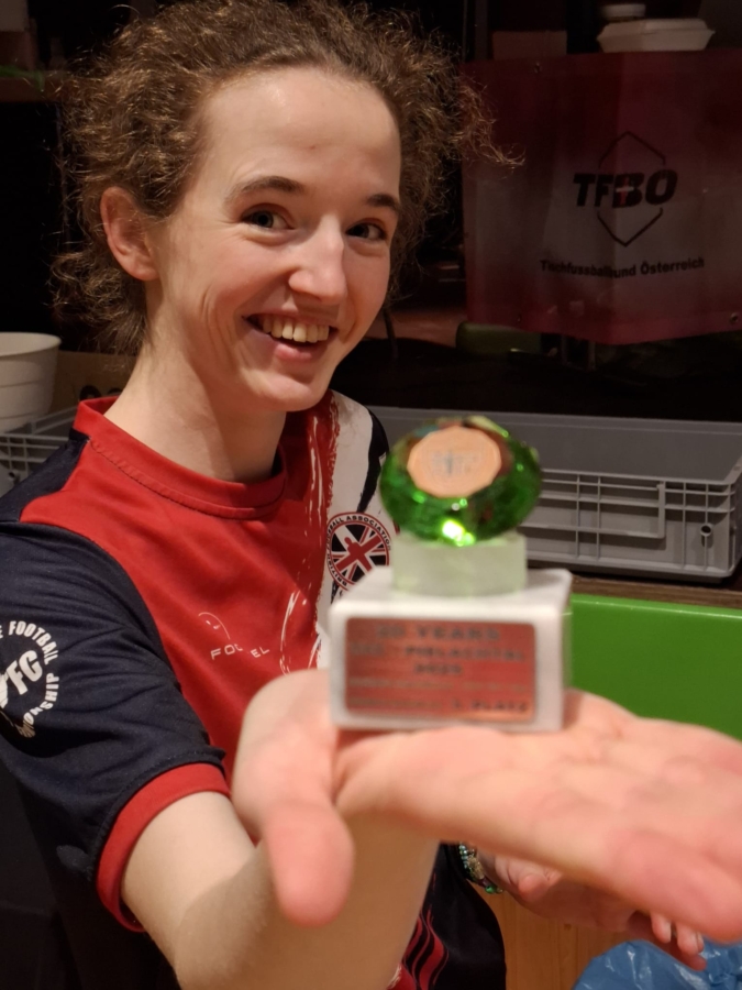 Eine lächelnde Person in einem roten und marineblauen Sporttrikot hält eine kleine grün-weiße Trophäe oder Auszeichnung in der ausgestreckten Hand, während im Hintergrund ein TFBO-Banner zu sehen ist.