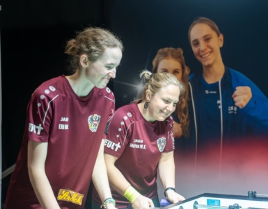 Zwei Frauen in kastanienbraunen Hemden stehen Seite an Seite und spielen Tischfußball, lächelnd und konzentriert. Ein großes Plakat im Hintergrund zeigt zwei weitere Frauen, von denen eine in Blau den Daumen nach oben streckt. Die Szene scheint bei einer Sportveranstaltung zu spielen.