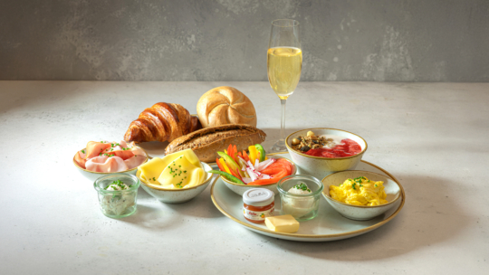 Ein Frühstücksbuffet mit Croissant, Brötchen, Käse, Schinken, Rührei, Butter, Gemüse, Müsli, Joghurt und einem Glas Sekt, angerichtet auf einem hellen Tisch.