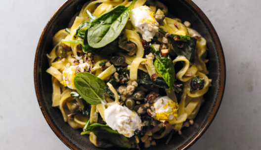 Eine Schüssel Nudeln mit frischem Blattspinat, cremigem Käse, Kapern, geriebener Zitronenschale und gehackten Nüssen, serviert in einer dunklen Schüssel auf einer hellen Unterlage.