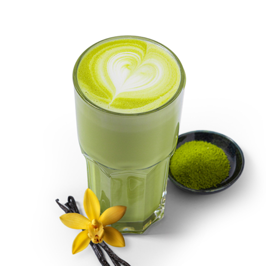 Ein Glas grüner Matcha Latte mit herzförmiger Latte Art obenauf, daneben eine kleine Schale mit Matcha-Pulver, Vanilleschoten und einer gelben Vanilleblüte, auf weißem Hintergrund.