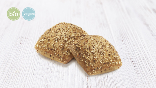 Zwei rechteckige Vollkornbrötchen mit Körnern belegt auf einer hellen Holzoberfläche. Auf den kreisförmigen Etiketten in der oberen linken Ecke steht "bio" und "vegan".