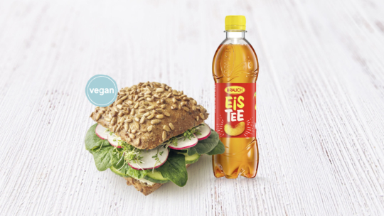 Ein Sonnenblumenkernsandwich mit Spinat, Radieschen und Sprossen, das als vegan gekennzeichnet ist, steht neben einer Flasche Rauch Eistee Pfirsich-Eistee auf einer weißen Holzfläche.