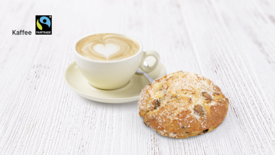 Eine Tasse Cappuccino mit herzförmigem Schaum sitzt auf einer Untertasse neben einem gezuckerten Gebäckstück auf einer weißen Holzfläche. Das Fairtrade-Logo und das Wort Kaffee erscheinen in der oberen linken Ecke.