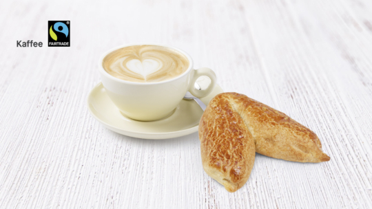 Eine Tasse Cappuccino mit herzförmigem Schaum sitzt auf einer Untertasse neben zwei goldbraunen Gebäckstücken auf einer weißen Holzfläche. Das Fairtrade-Logo und das Wort Kaffee erscheinen in der oberen linken Ecke.