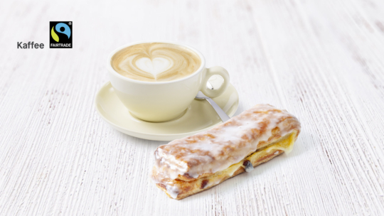 Eine Tasse Kaffee mit herzförmigem Milchkaffee steht auf einer Untertasse mit einem Löffel neben einem glasierten Gebäck auf einer weißen Holzfläche. Das Fairtrade-Kaffee-Logo und das Wort Kaffee befinden sich in der oberen linken Ecke.